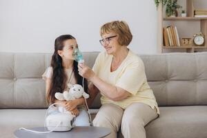 Little girl making inhalation with nebulizer with granny. Woman makes inhalation to a sick child while embracing her. Grandmother helping granddaughter with cold and flu to inahale nebuliser aerosol. photo