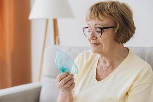 Sick senior woman has a viral infection or flu, labored breathing, asthma. An elderly female is using inhaler, nebulizer for self treatment sitting alone on the sofa at home photo