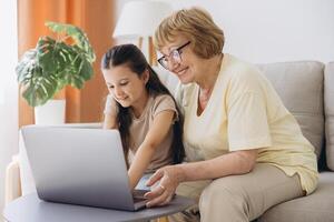 Multi generational family, grandma and granddaughter sit on couch with wireless computer, look at each other with tenderness and love, enjoy leisure on internet and time together, watch on-line movies photo