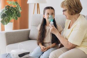 Little girl making inhalation with nebulizer with granny. Woman makes inhalation to a sick child while embracing her. Grandmother helping granddaughter with cold and flu to inahale nebuliser aerosol. photo