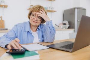 Shocked puzzled senior woman reading paper document, reviewing agreement terms, checking bills for payment at home table, getting angry about mistake, too high price, problem photo