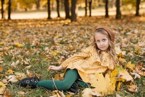 Portrait of a beautiful little blonde girl in a yellow dress in an autumn park lying in the middle of maple leaves. photo
