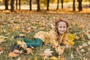 Portrait of a beautiful little blonde girl in a yellow dress in an autumn park lying in the middle of maple leaves. photo