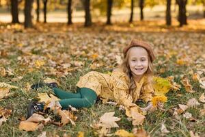 Portrait of a beautiful little blonde girl in a yellow dress in an autumn park lying in the middle of maple leaves. photo