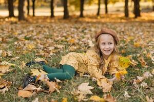 Portrait of a beautiful little blonde girl in a yellow dress in an autumn park lying in the middle of maple leaves. photo