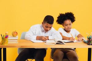 Happy african american schoolgirl and mixed race schoolboy sitting together at desk and studying on yellow background. Back to school concept. photo