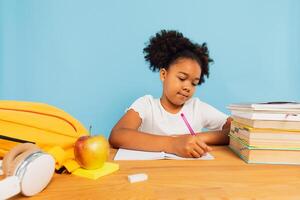 Happy African American schoolgirl doing homework at desk in class on blue background. Back to school concept. photo