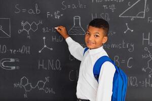 Happy mixed race schoolboy solving problems near the blackboard at school, back to school concept. photo