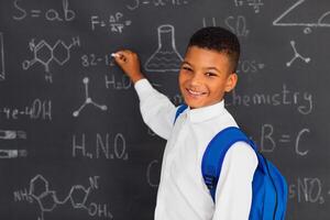 Happy mixed race schoolboy solving problems near the blackboard at school, back to school concept. photo