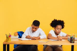 Happy african american schoolgirl and mixed race schoolboy sitting together at desk and studying on yellow background. Back to school concept. photo