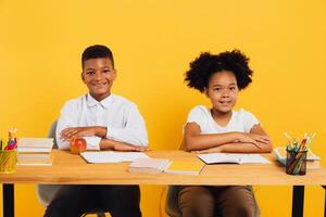 Happy african american schoolgirl and schoolboy sitting together at desk and studying on yellow background. Back to school concept. photo