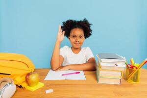 Happy African American schoolgirl sitting at desk in class and stretching hand uphill on blue background. Back to school concept. photo