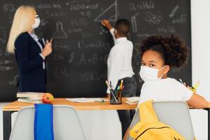 Portrait of African American school girl in mask sitting at desk on a blackboard background in a classroom during a lesson. Mask mode, quarantine, covid-19. photo