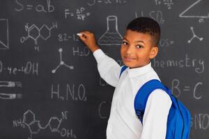 Happy african schoolboy solving problems near the blackboard at school, back to school concept photo