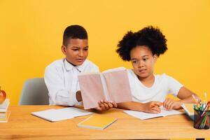 Happy african american schoolgirl and mixed race schoolboy sitting together at desk and studying on yellow background. Back to school concept. photo