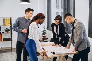 Group of young male and female students sharing ideas while working on project correcting mistakes in architectural drawings without using technologies collaborating in coworking space with bid desk photo
