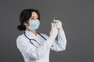 Female doctor with stethoscope in mask holding injection, antidote against coronavirus on gray background. photo