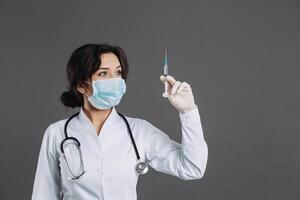 Female doctor with stethoscope in mask holding injection, antidote against coronavirus on gray background. photo