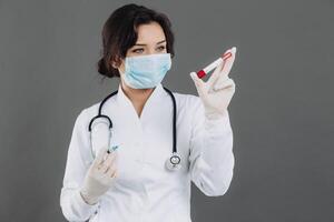 Female doctor with stethoscope in mask holding injection, antidote against coronavirus on gray background. photo
