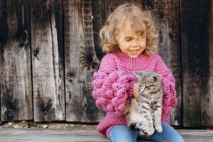 Portrait of a beautiful and happy curly girl in a knitted sweater playing with a kitten near a wooden wall photo