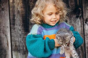 Portrait of a beautiful and happy curly girl in a knitted sweater playing with a kitten near a wooden wall photo