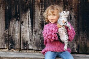 Portrait of a beautiful and happy curly girl in a knitted sweater playing with a kitten near a wooden wall photo