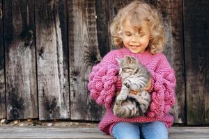 Portrait of a beautiful and happy curly girl in a knitted sweater playing with a kitten near a wooden wall photo