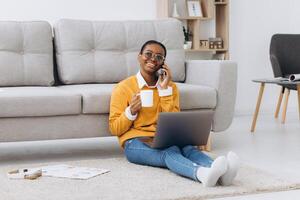 Beautiful young black university student sitting on the floor drinking coffee, talking on the phone and doing a project on a laptop photo
