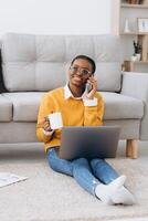 Beautiful young black university student sitting on the floor drinking coffee, talking on the phone and doing a project on a laptop photo