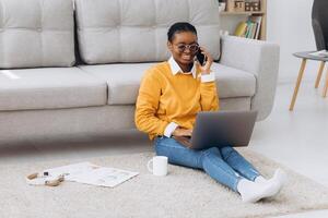 Beautiful young black university student sitting on the floor drinking coffee, talking on the phone and doing a project on a laptop photo