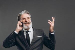 Business and fashionable bearded mature man in a gray suit smiling, talking by phone and showing copy space on the grey background. photo