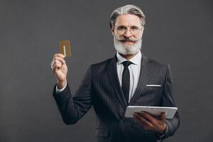 Business and fashionable bearded mature man in a gray suit making shopping in tablet with his gold credit card on the grey background. photo