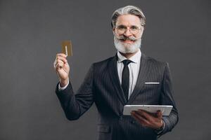 Business and fashionable bearded mature man in a gray suit making shopping in tablet with his gold credit card on the grey background. photo