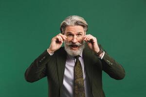 Bearded middle age teacher in khaki suit holds glasses with his hands and blinks his eyes with fun on green background photo