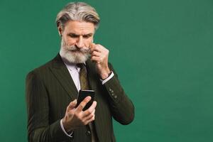 Stylish middle-aged man in khaki suit looks at phone and fixes mustache and beard on green background photo