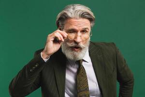Bearded middle age teacher in khaki suit holds glasses with his hands and blinks his eyes with fun on green background photo
