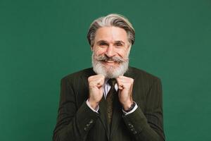 A middle-aged bearded man in a suit holds his fists, aches and rejoices on a green background. photo