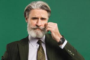 Stylish middle-aged man in khaki suit fixes mustache and beard on green background photo