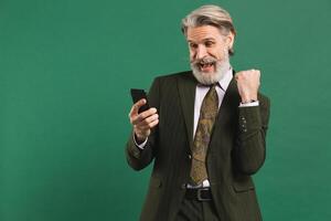 Bearded middle-aged man in khaki suit screaming loudly and looking at phone while holding fist of happiness against green background photo