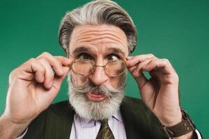 Bearded middle age teacher in khaki suit holds glasses with his hands and blinks his eyes with fun on green background photo