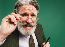 Bearded and mustachioed middle-aged teacher in khaki suit holds hand with glasses and looks close up and says something on green background photo