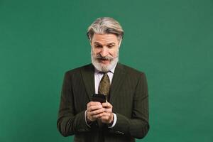 Bearded middle-aged man in khaki suit looks at phone and shows shock on green background photo