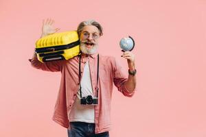 Happy bearded man in sunglasses with a camera holds a suitcase and a globe and prepares for the trip on a pink background. photo