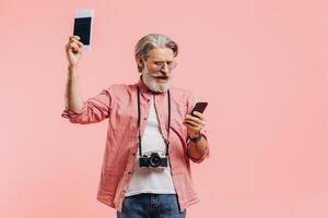 Happy bearded man in sunglasses holding a mobile phone and a passport with tickets on the pink background photo