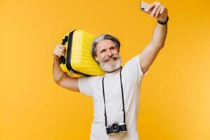 Stylish bearded and mustachioed man smiling and taking a selfie on his smartphone with a yellow suitcase. photo