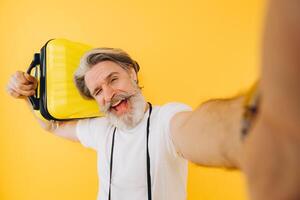 Stylish bearded and mustachioed man smiling and taking a selfie on his smartphone with a yellow suitcase. photo