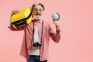Happy bearded man in sunglasses with a camera holds a suitcase and a globe and prepares for the trip on a pink background. photo