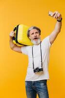 Stylish bearded and mustachioed man smiling and taking a selfie on his smartphone with a yellow suitcase. photo