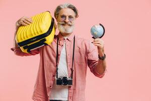 Happy bearded man in sunglasses with a camera holds a suitcase and a globe and prepares for the trip on a pink background. photo