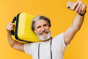 Stylish bearded and mustachioed man smiling and taking a selfie on his smartphone with a yellow suitcase. photo